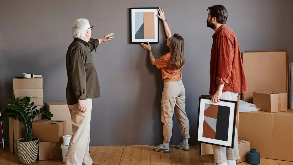 Horizontal long shot of young man and woman helping grandpa to decorate living room in new house