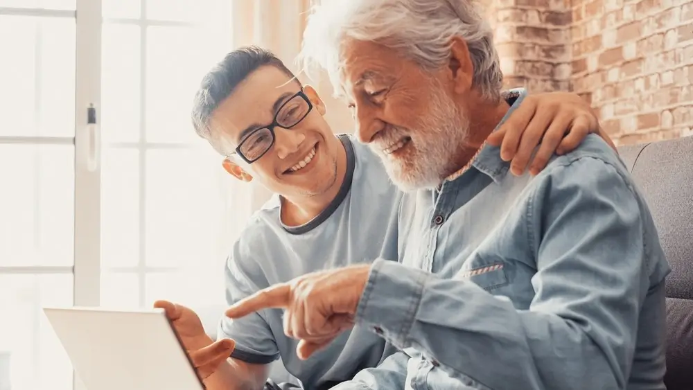 Smiling young boy helping senior grandfather with laptop choose goods or services via internet or web surfing together at home. Younger generation caring about older relatives teaching using computer