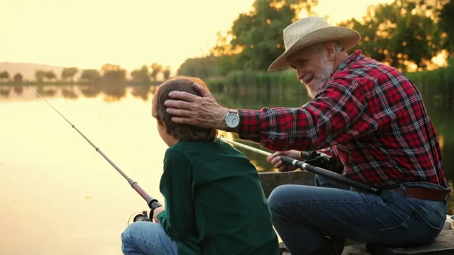 Back view of the senior man chatting with his grandson and stocking him at the head while sitting with the fishing roads and fishing at the pier.