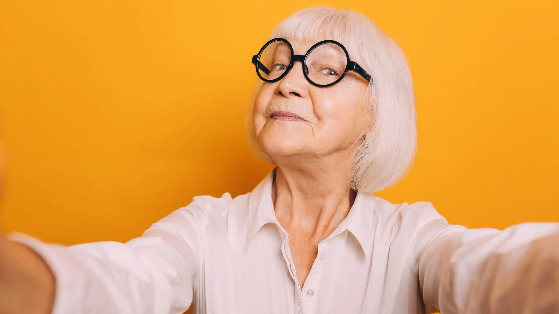 Portrait of elderly woman with short white hair wearing white glasses and light blouse holding camera and taking selfie. Old Woman isolated over orange background