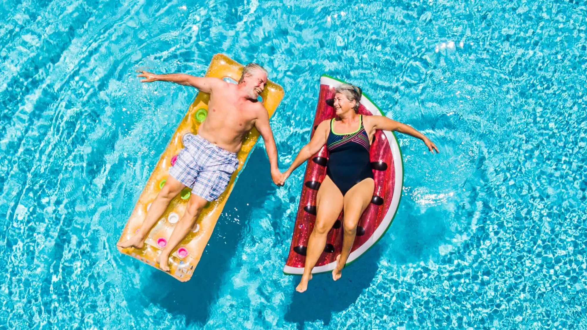 Above vertical view of people old senior couple taking hands with love and having fun on the blue clear swimming pool together enjoying the summer holiday vacation with trendy coloured lilos mattress