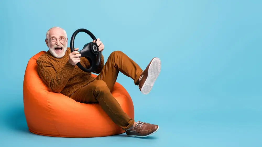 Portrait of his he nice attractive cheerful cheery glad funny bearded grey-haired man sitting in bag chair having fun holding in hands steering wheel isolated over blue pastel color background