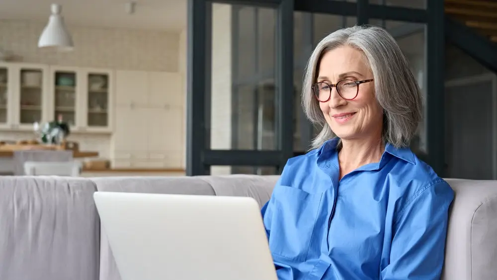 Older smiling 60s woman sitting at home on sofa, holding laptop. Senior happy business woman using pc technologies, working, chatting, spending time in social media internet, ecommerce.