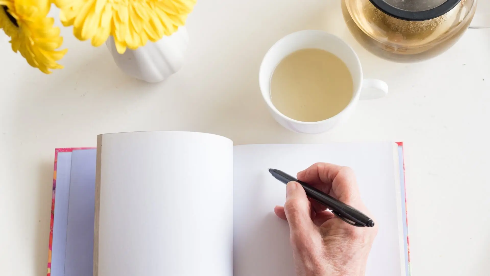 High angle view of older woman's hand holding black pen on blank journal with cup of chamomile tea and yellow flowers (selective focus)