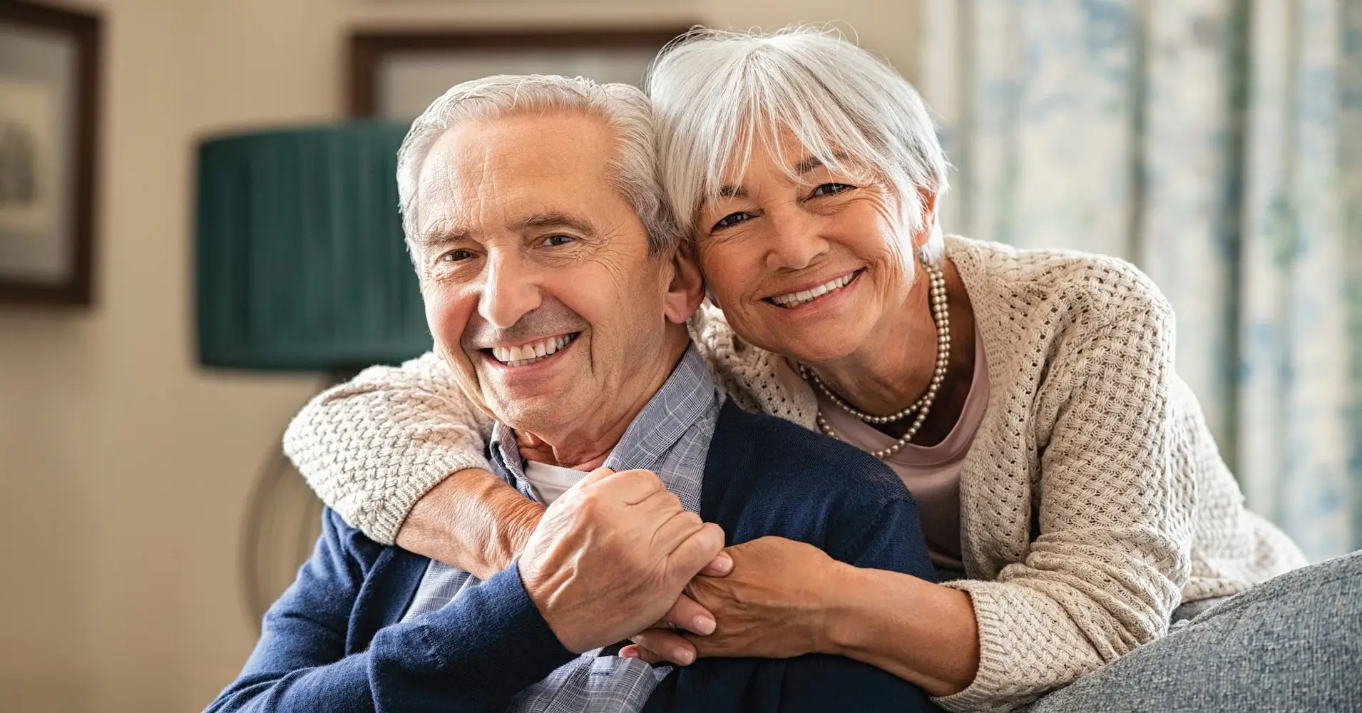 Portrait of romantic senior man with his beautiful wife stay at home. Smiling and caring old woman embracing from behind her retired husband sitting on couch. Cheerful old couple looking at camera.