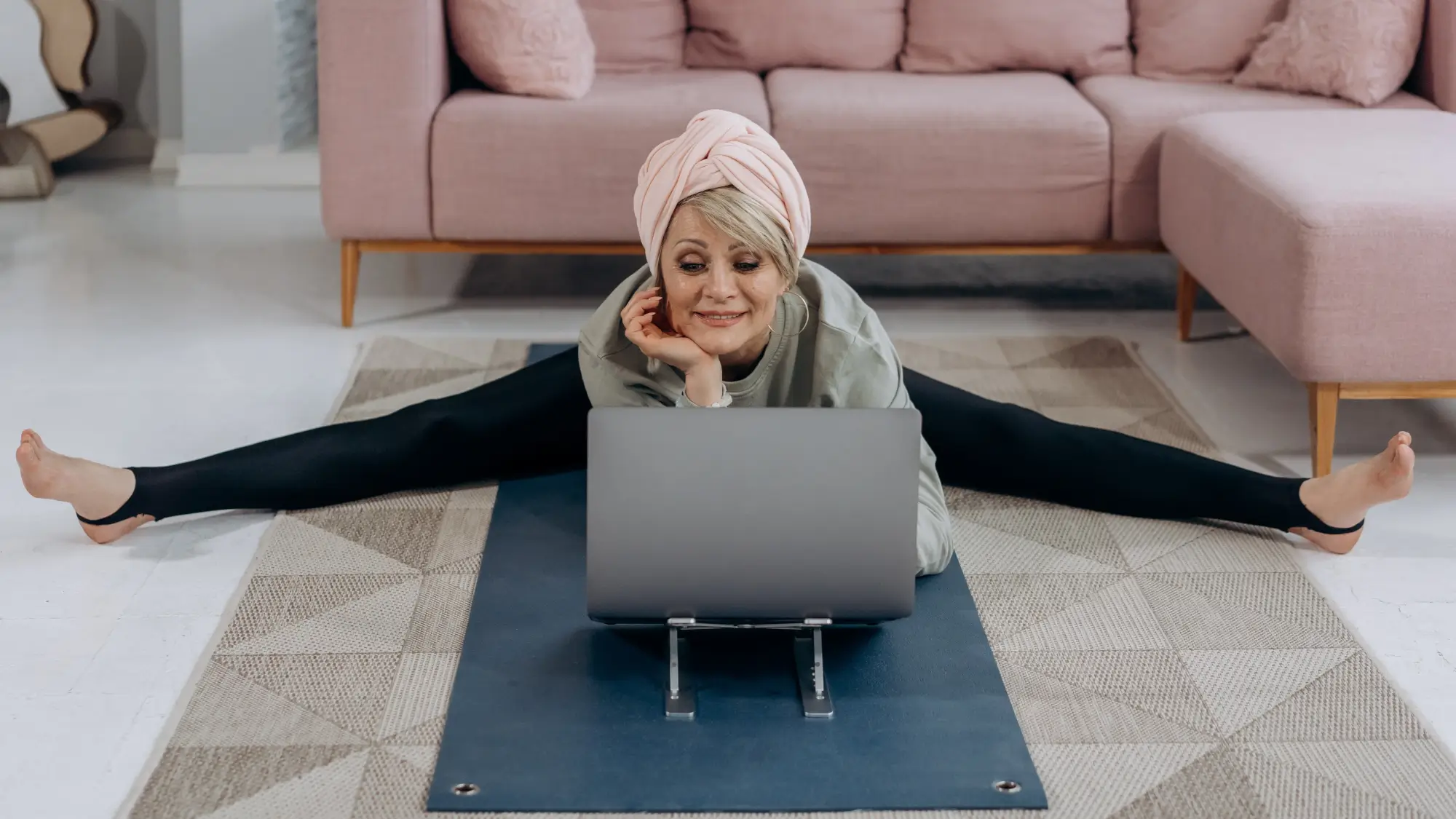 Old woman stretching on yoga mat in her living room while working on her laptop