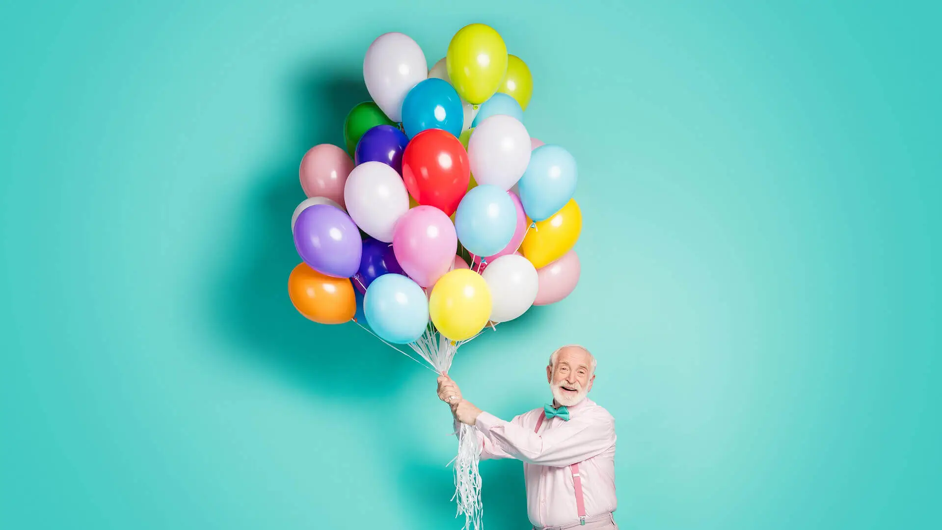 Portrait of positive cheerful old man hold many helium balloons fly air sky enjoy festive occasion wear pink trousers isolated over teal color background