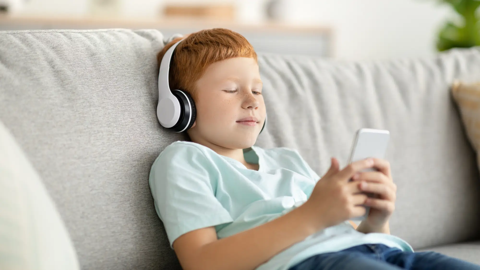 Redhead preteen boy sitting on couch, watching videos or listening to music on mobile phone, using wireless headset, copy space. Modern technologies and kids entertainment concept