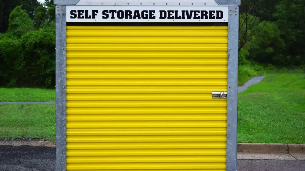 A self storage pod or shed with a yellow door and lock. Sign indicates a deliverable self-storage unit. Unit is on a street or parking lot with green grass and trees in background. Outdoors in daytime