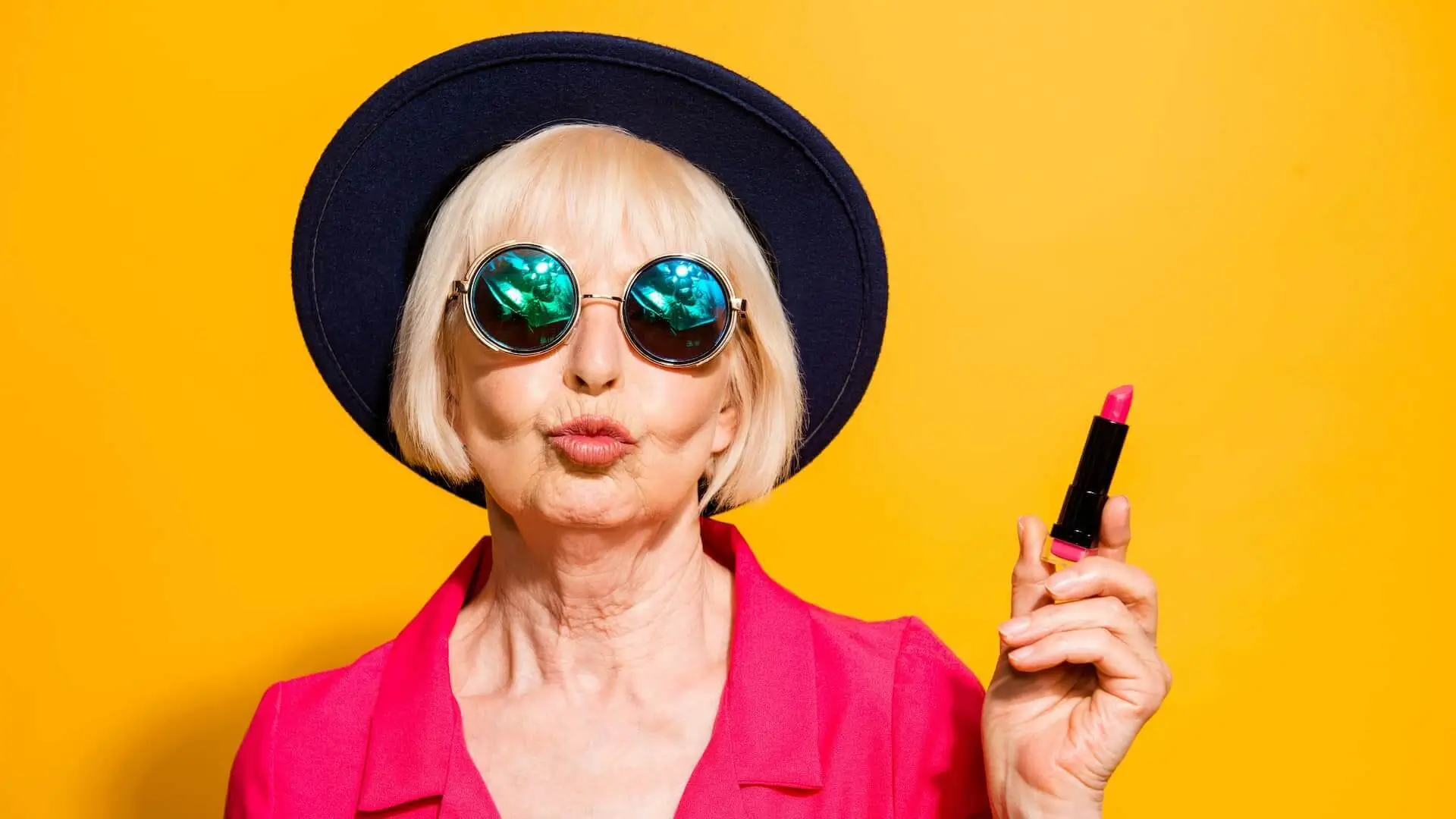 Grandmother getting ready for date! Charming, attractive old woman holding red lipstick pouting lips for a kiss isolated on bright yellow background.