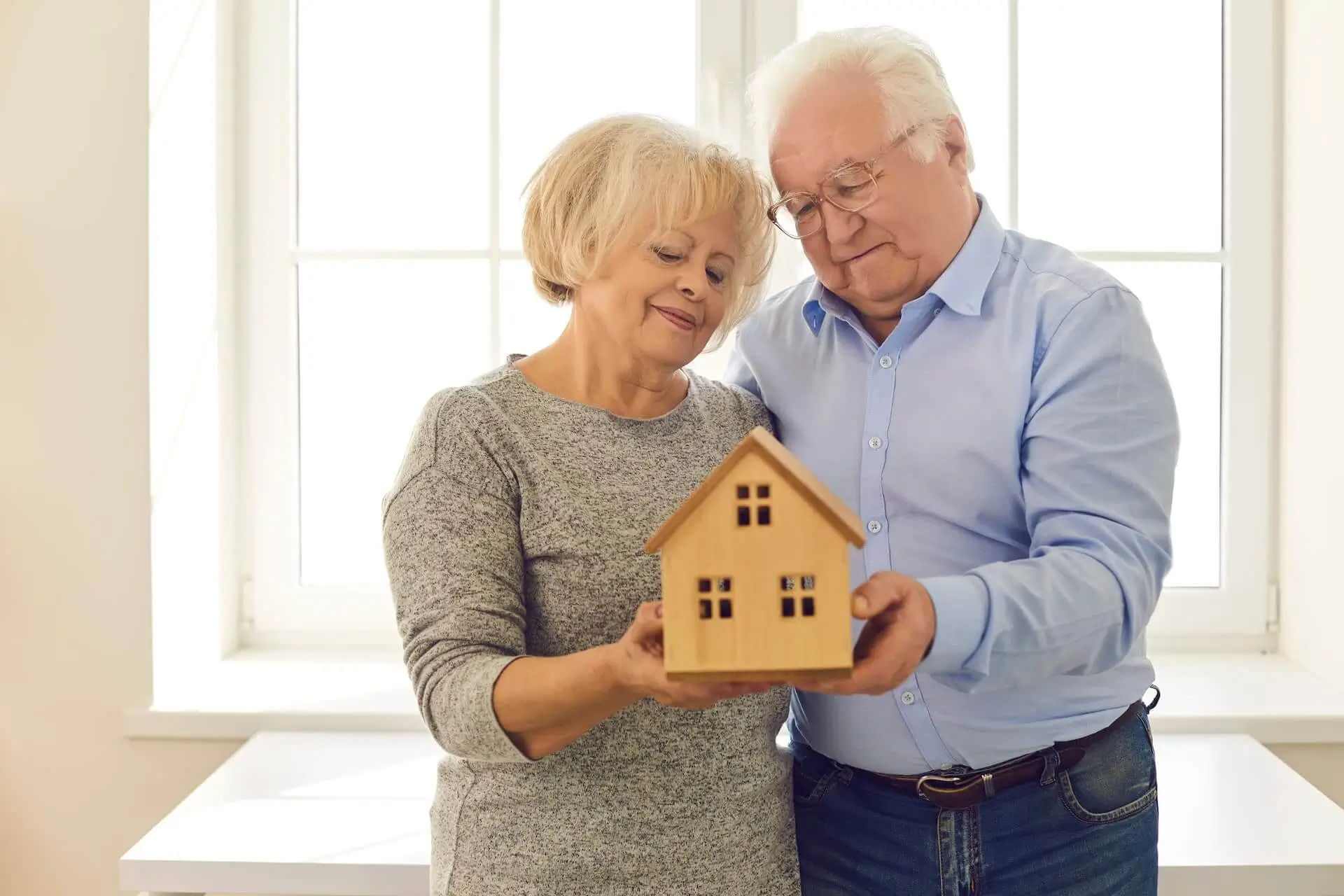Smiling senior couple looking at a wooden model of a house being held in their hands standing by window in new home. Portrait of retirees who have purchased property insurance. New house concept.