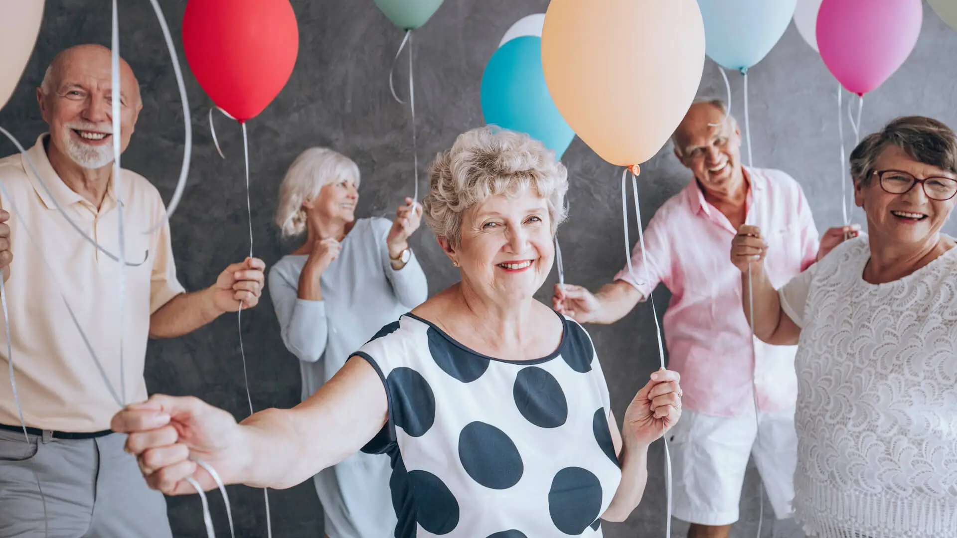 Smiling grandmother wearing a blouse with black dots during New Year's Eve party with friends holding colorful balloons