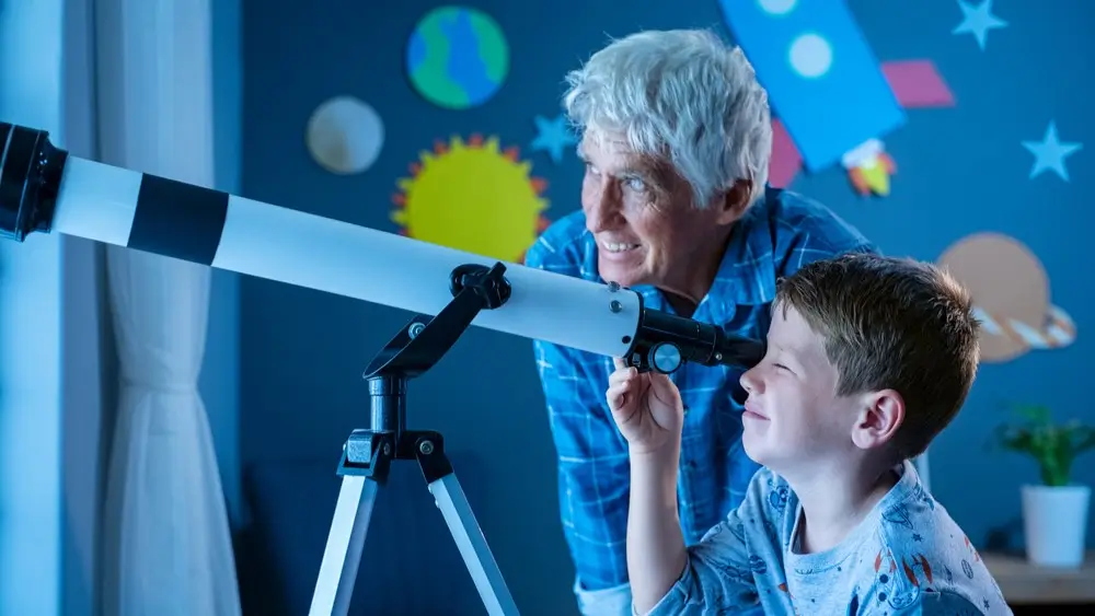 Grandfather teaching grandson using telescope to see planets and galaxy. Child watching stars through a telescope at night with senior man. Grandpa and grandchild looking together at positive future.